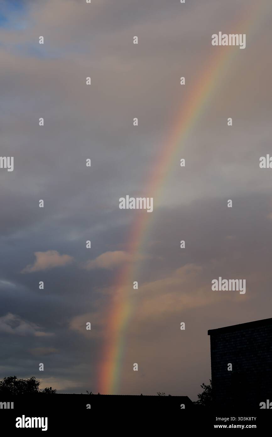 Abendliche Regenbogenbögen über Sturmwolken und Dächern in Amsterdam, Niederlande, von einem Wohnviertel unter einem violetten Abendhimmel aus gesehen. Stockfoto