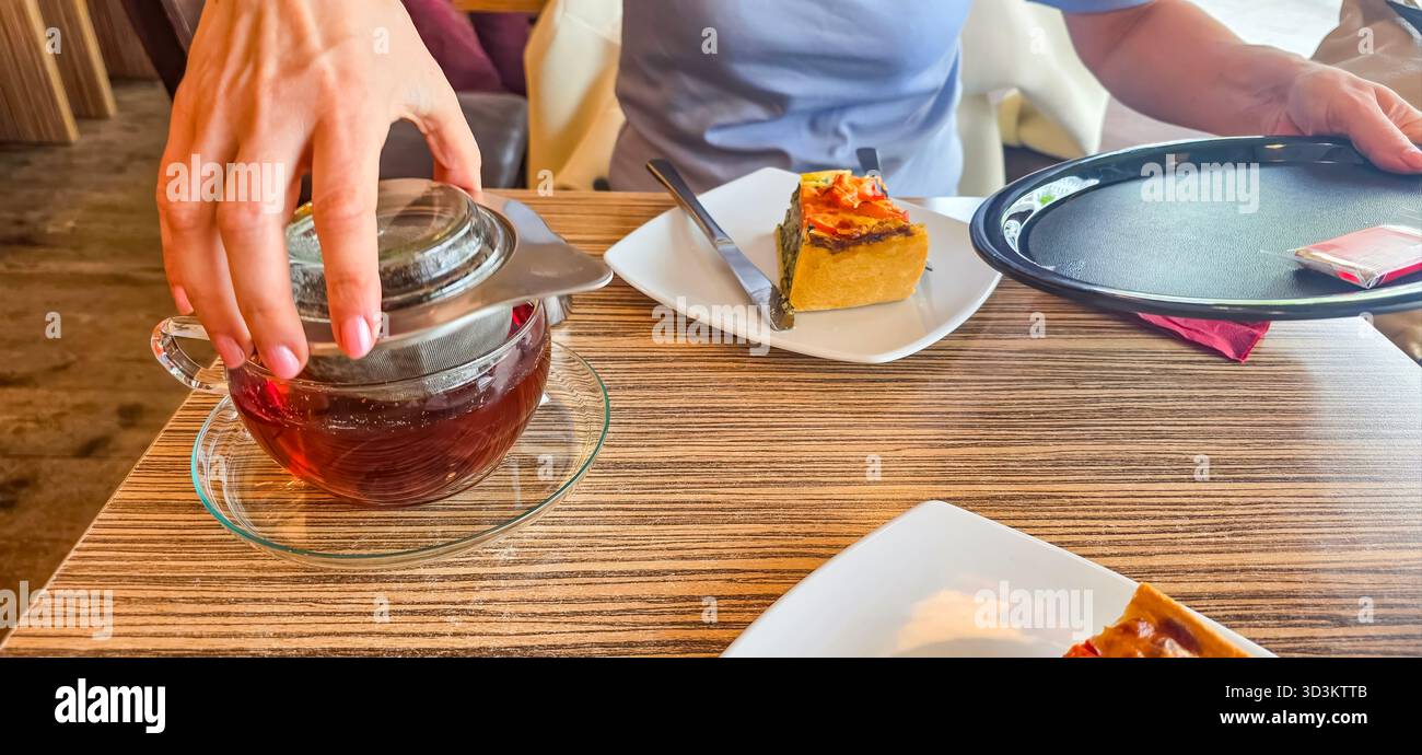 Hand gießt Tee aus einer Teekanne aus Glas auf einen Holztisch, begleitet von einem Stück Dessert und einem Teller, was eine gemütliche Atmosphäre schafft. - Smartphone-aufgenommenes Stockfoto