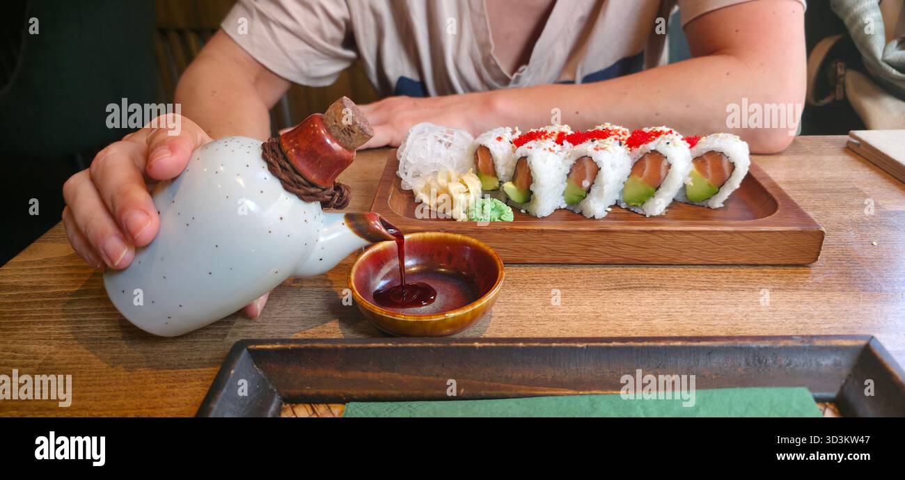 Hand gießt Sojasauce über Sushi-Brötchen auf einem Holztisch, begleitet von Wasabi und Ingwer, was ein lebhaftes kulinarisches Erlebnis schafft. - Smartphone-aufgenommenes Stockfoto