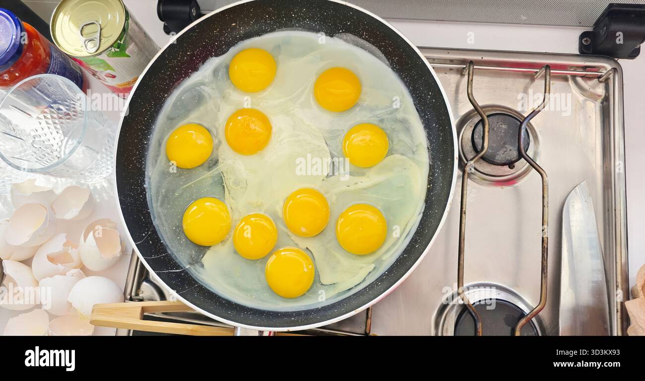 . Frische Eier werden in einer Pfanne auf einem Herd gebraten, umgeben von Küchenutensilien und leeren Eierschalen, wodurch eine lebendige Kochszene entsteht - Smartphone-aufgenommenes Stockfoto