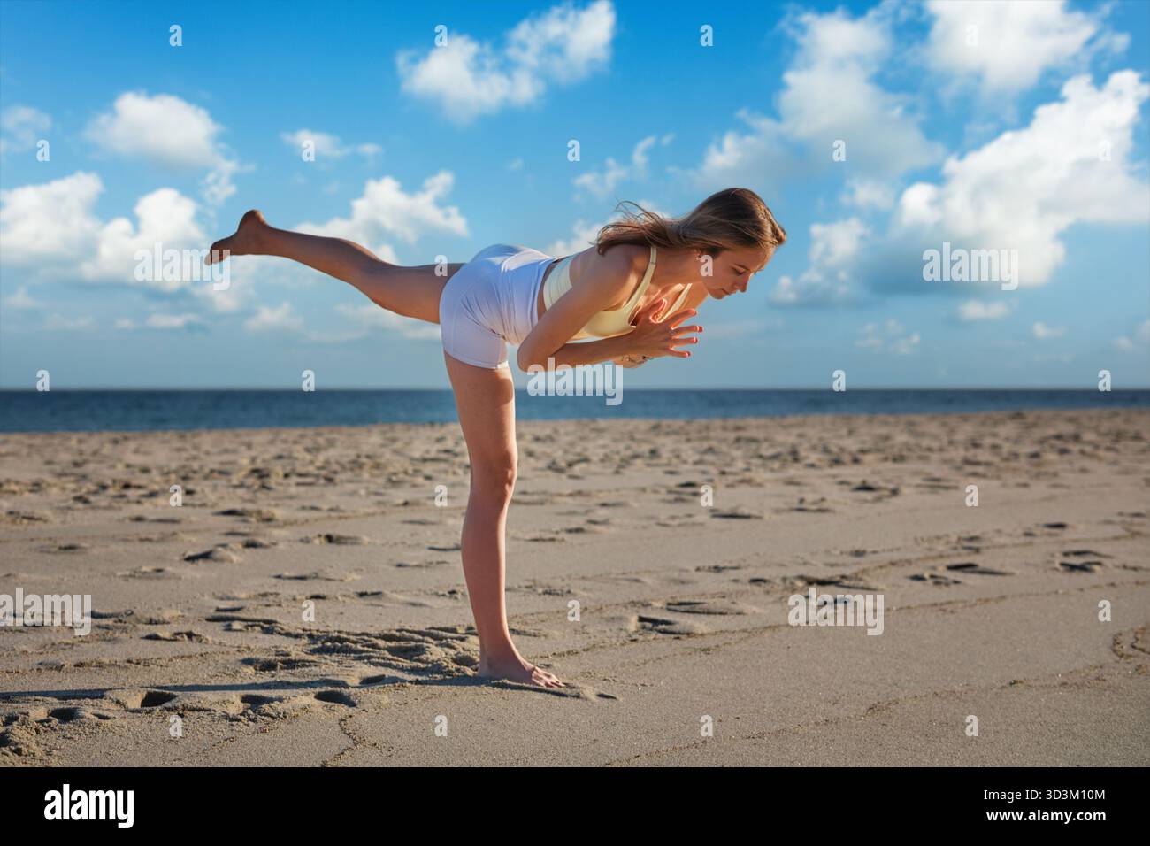Eine Frau hält anmutig die Virabhadrasana Warrior 3 Pose am Strand und balanciert auf einem Bein. Die Sonne geht im Hintergrund unter und wirft warme Farben auf Stockfoto