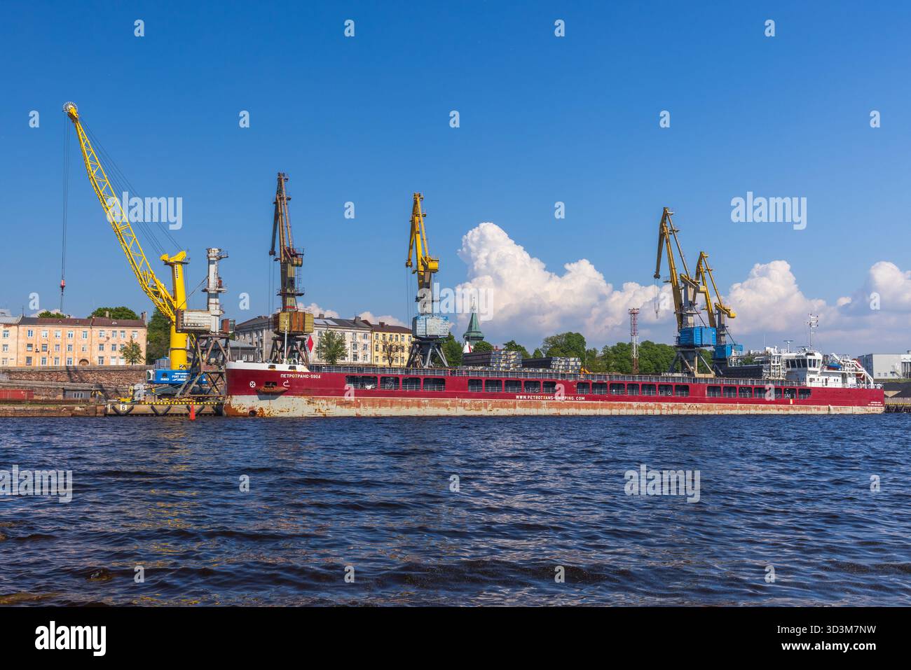 Vyborg, Russland - 27. Mai 2024: Eine lebhafte Hafenszene zeigt ein langes rotes Frachtschiff, das neben aufragenden gelben Kranichen, ruhigem Wasser und einer klaren blauen Ske vor Anker liegt Stockfoto
