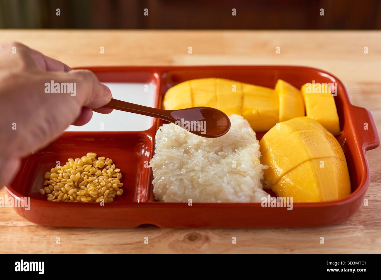 Eine Hand greift nach einem Löffel Sirup, der bereit ist, über eine köstliche Portion süßen Kleberreis, frische Mangoblätter und knusprige Mungbohnen hineinzuträufeln Stockfoto