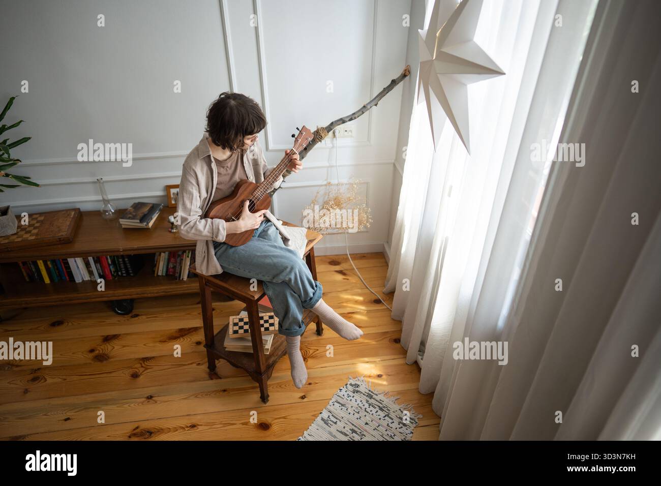 Junge Frau spielt Melodie auf hölzerner Ukulele-Gitarre, sitzt vor dem Fenster im gemütlichen Wohnzimmer. Stockfoto