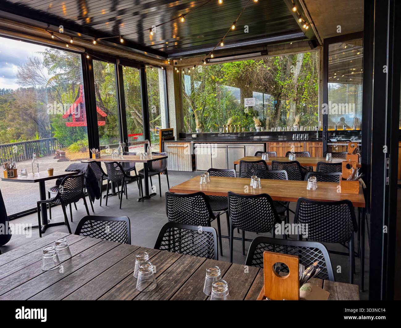 Pataka Kai Restaurant in Rotorua, Neuseeland. Blick auf den leeren Speisesaal mit Holztischen, schwarzen Stühlen und Lichterketten, mit Blick auf üppig Stockfoto