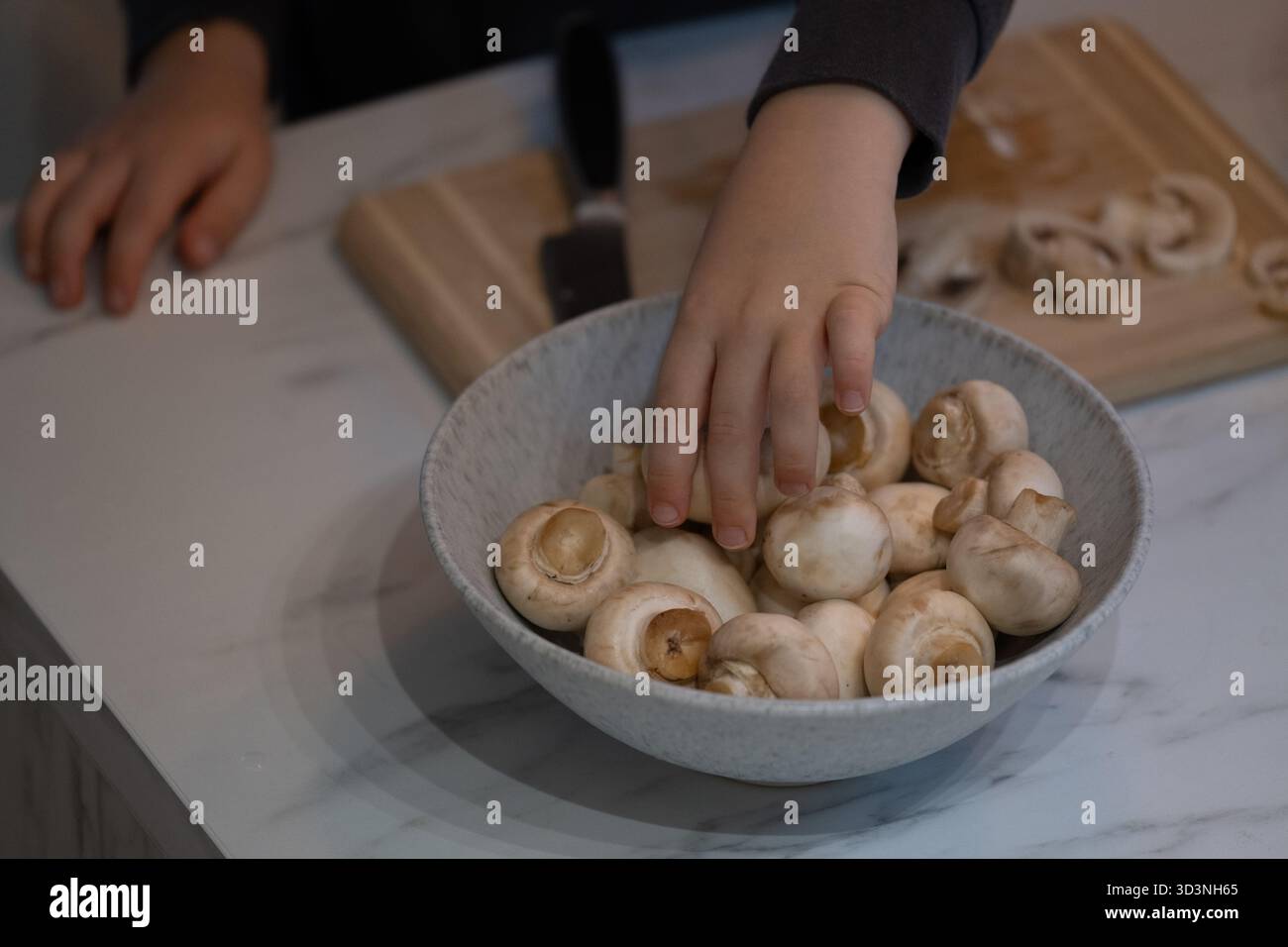 Die Hand eines Kindes pflückt während der Hausmannskost frische Pilze aus einer Schüssel. Diese Close-up-Küche Szene repräsentiert Familienzeit, gesunde Ernährung, Lebensmittelzubereitung Stockfoto