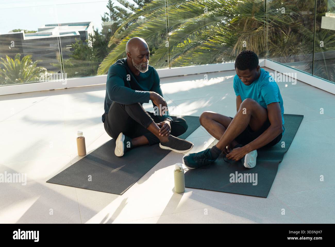 Sitzen afroamerikanischer Vater und Sohn, die Schuhe auf der sonnigen Terrasse anpassen, mit Auflagen, Wasserflaschen Stockfoto