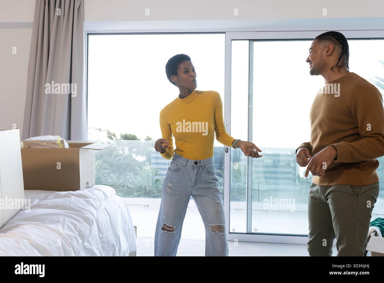 Lächelnde, abwechslungsreiche Paare tanzen im modernen Apartment-Schlafzimmer mit Umzugskasten und Balkon-Blick Stockfoto