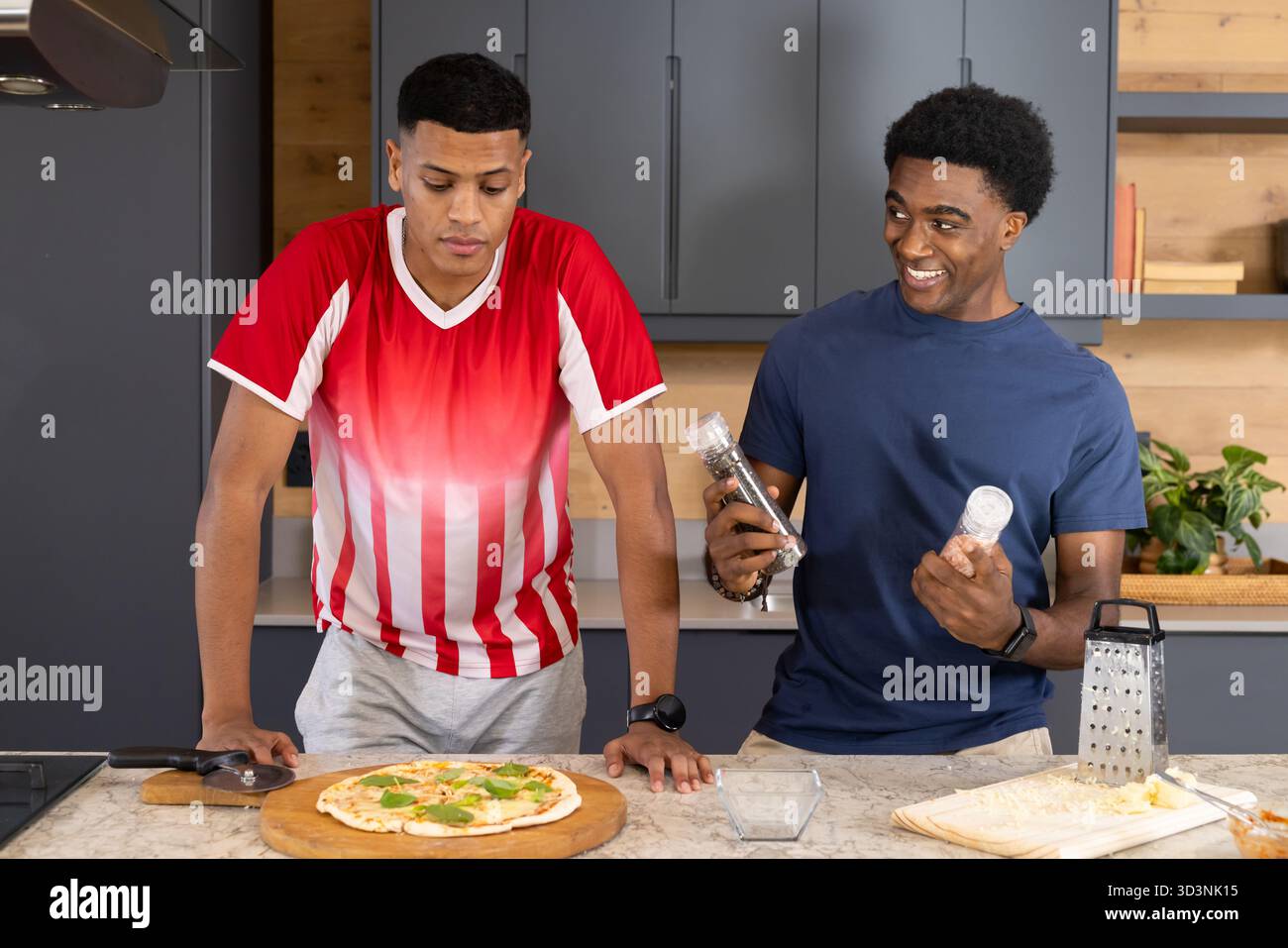 Verschiedene männliche Freunde würzen Pizza auf Marmor-Arbeitsplatte in der Küche, mit Salz- und Pfeffermühle Stockfoto