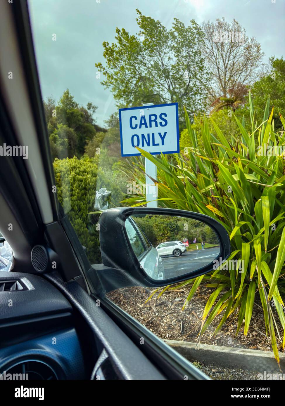 Das Parkschild „nur Fahrzeuge“ wird an einem Pfosten angebracht, aus dem Fahrzeuginnenraum gesehen und ein Seitenspiegel reflektiert ein anderes Fahrzeug. Grünes Laub und eine bewölkte Stockfoto