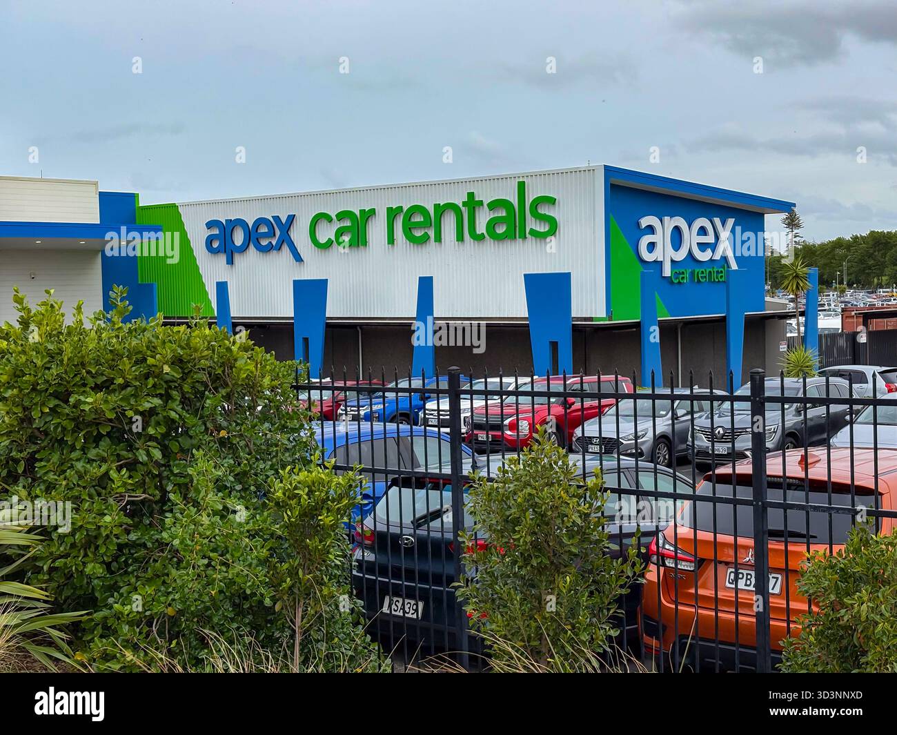 APEX Car Rentals Außengebäude mit geparkten Autos, sichtbar hinter einem Zaun und Laub am Flughafen Auckland, Neuseeland. Steht für Reise und Transaktion Stockfoto