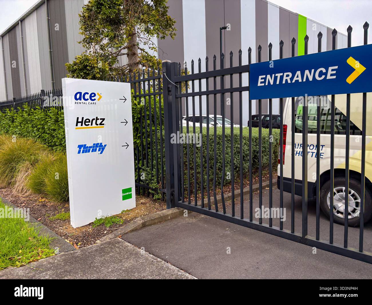 Schilder für Ace Rental Cars, Hertz und Thrifty am Eingang zu einer Autovermietung an einem Flughafen in Neuseeland. Ein Flughafen-Shuttle-Van ist sichtbar. Stockfoto