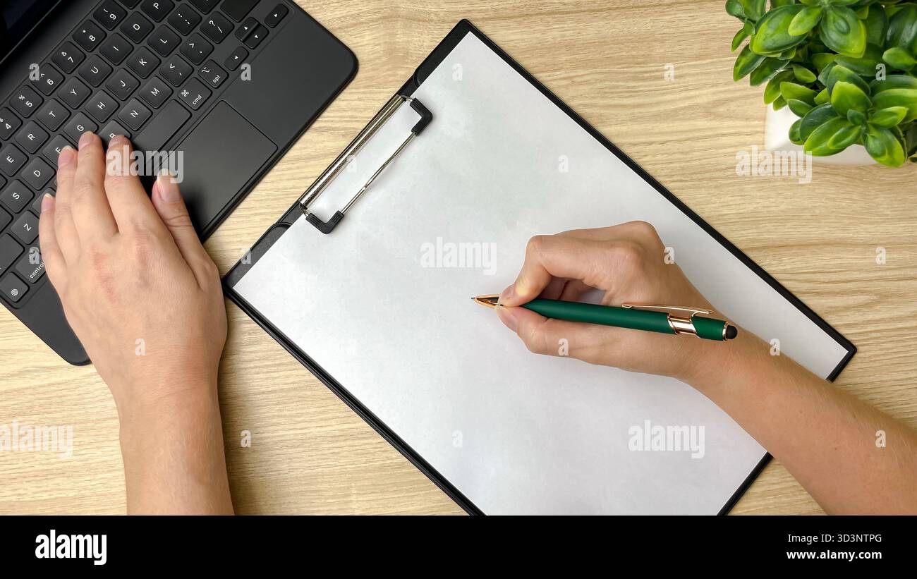 Die Hände einer Person über dem Laptop – eine auf der Tastatur, die andere bereit, Notizen zu machen. - Smartphone-aufgenommenes Stockfoto