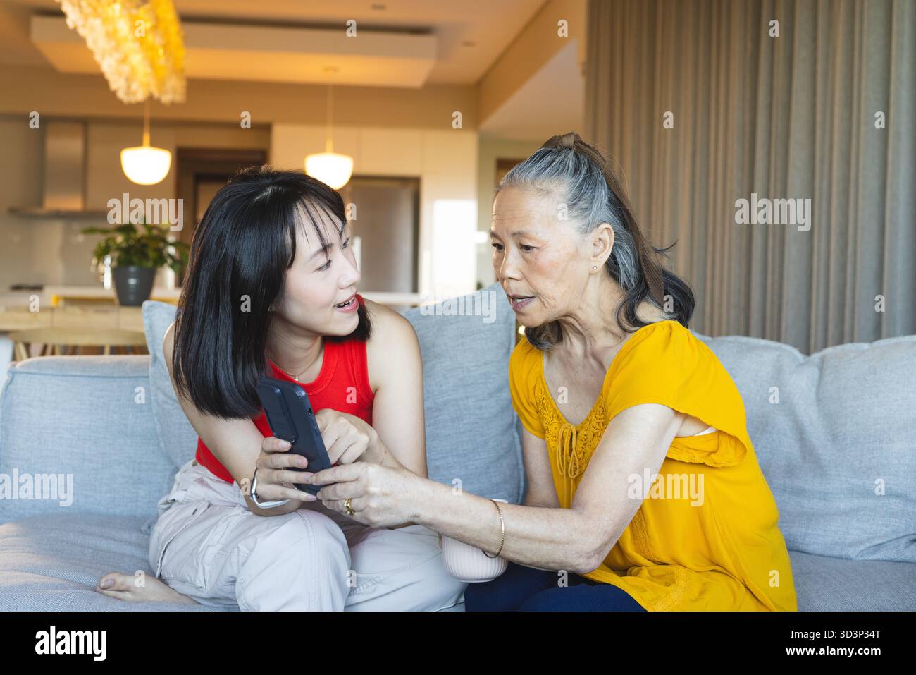 Asiatische Mutter und Tochter auf hellgrauem Sofa im modernen Wohnzimmer mit Smartphone Stockfoto