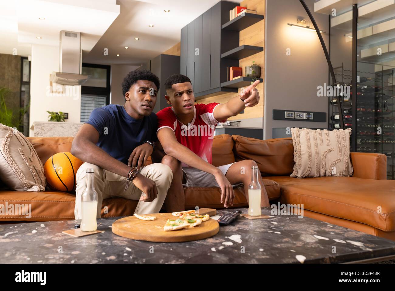 Verschiedene männliche Freunde, die sich nach vorne lehnen und in einem modernen, offenen Wohnzimmer zeigen, mit Pizzabett Stockfoto