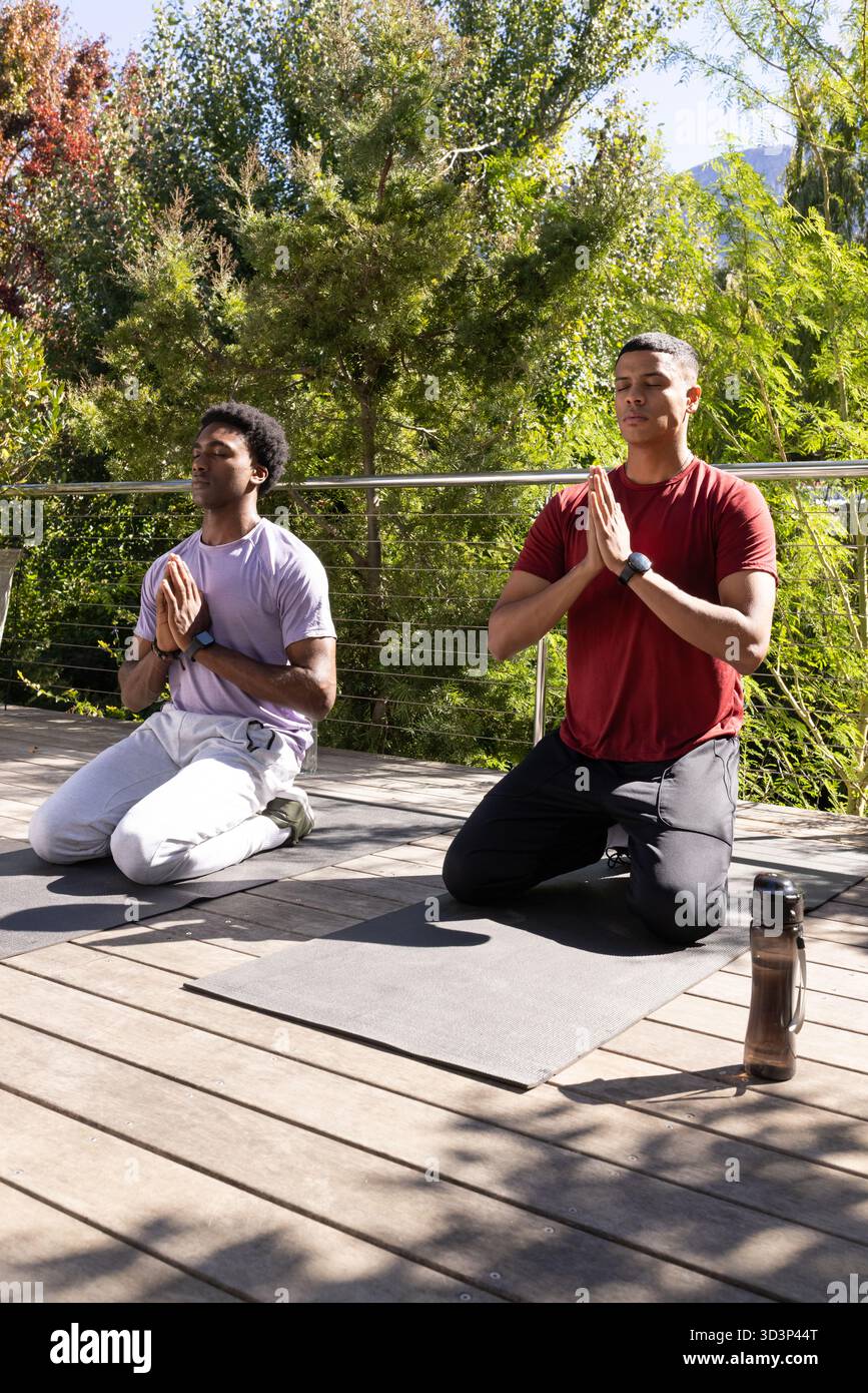Kniende männliche Freunde, die auf Yogamatten auf der Terrasse meditieren, mit einer Wasserflasche Stockfoto