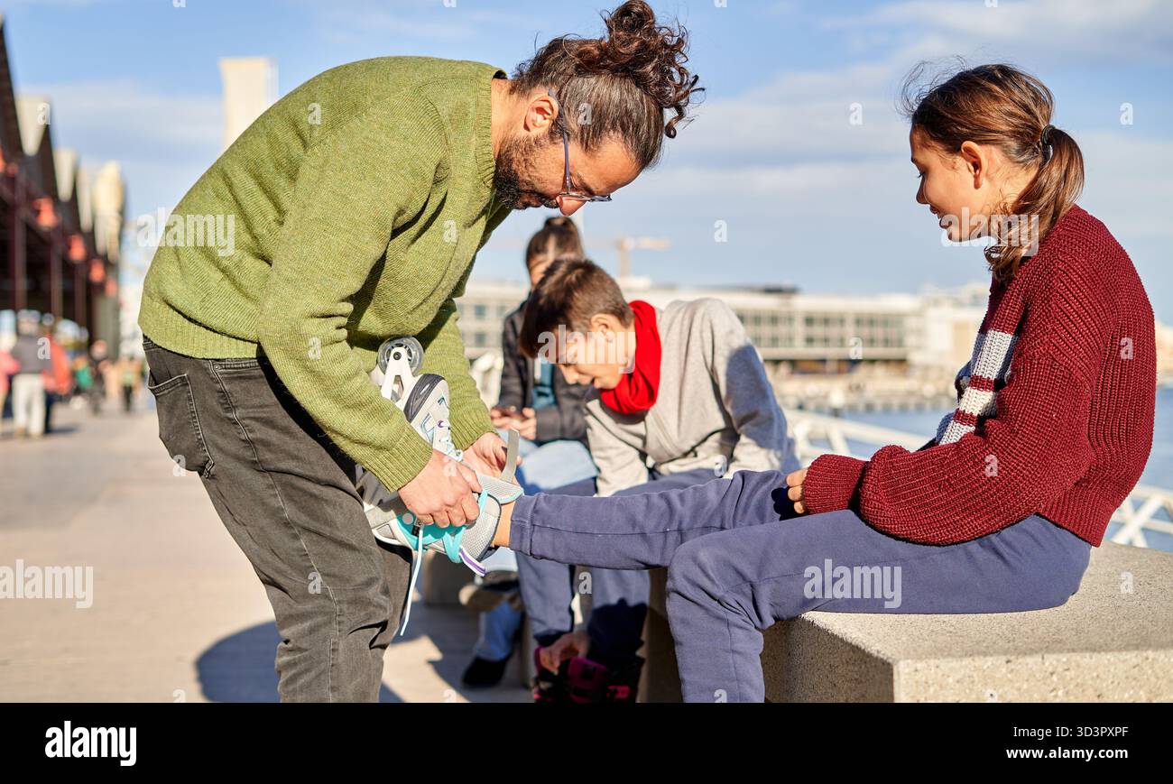 Ein Mann hilft einem Mädchen, ihre Schuhe anzuziehen, während zwei Jungs in der Nähe sitzen. Sie sind an einem sonnigen Tag in der Nähe des Ufers bereit für lustige gemeinsame Aktivitäten. Stockfoto
