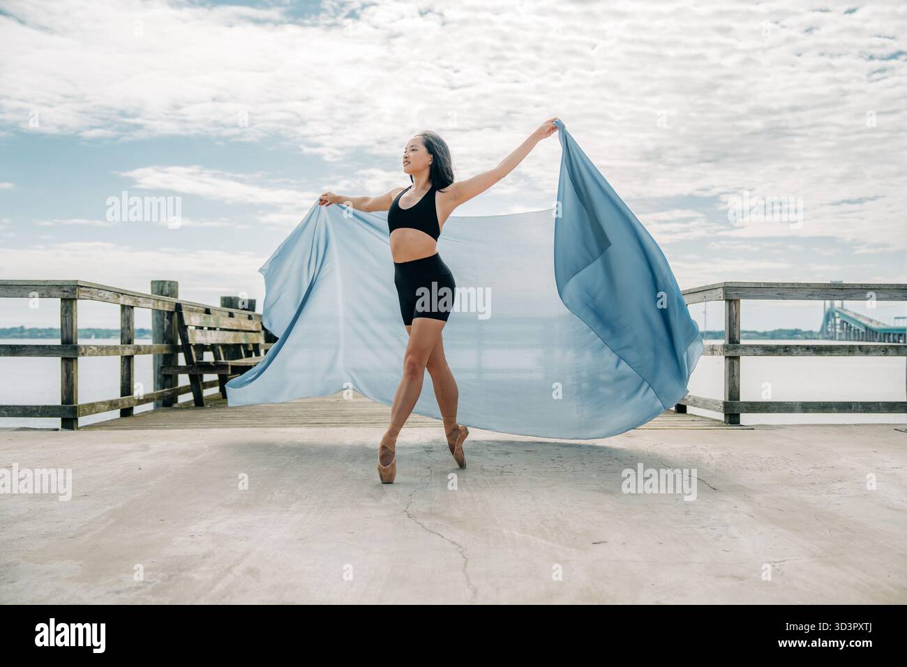 Balletttänzer streckt die Arme mit blauem Stoff am Pier am Wasser aus Stockfoto
