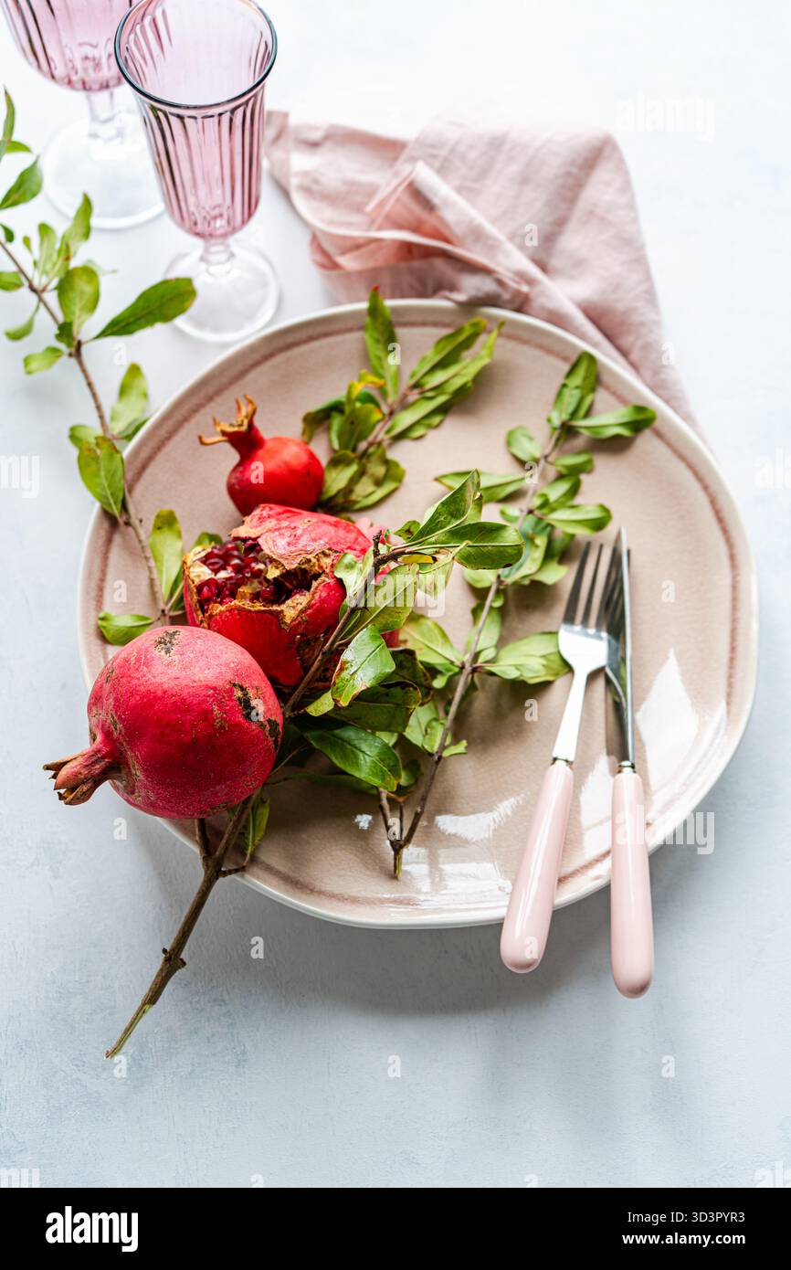 Von oben hält eine beige Keramikplatte einen Granatapfel mit grünen Blättern. Besteck mit rosafarbenem Griff und pinkfarbene Gläser ergänzen eine weiche rosafarbene Serviette auf einem li Stockfoto