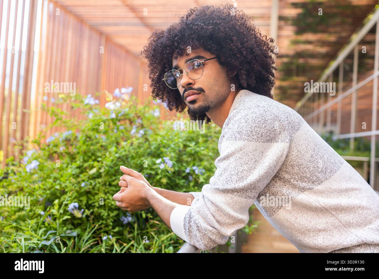 Ein junger Mann mit lockigen Haaren und Brillen lehnt sich über ein Geländer, umgeben von Pflanzen in einem städtischen Garten. Er strahlt ein entspanntes und nachdenkliches Verhalten aus, e Stockfoto