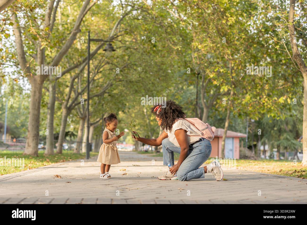 Afroamerikanische Mutter und Tochter genießen einen verspielten Moment in einem grünen Park. Die Mutter kniet und präsentiert einen Zweig, während das Kleinkind aufgeregt angreift Stockfoto