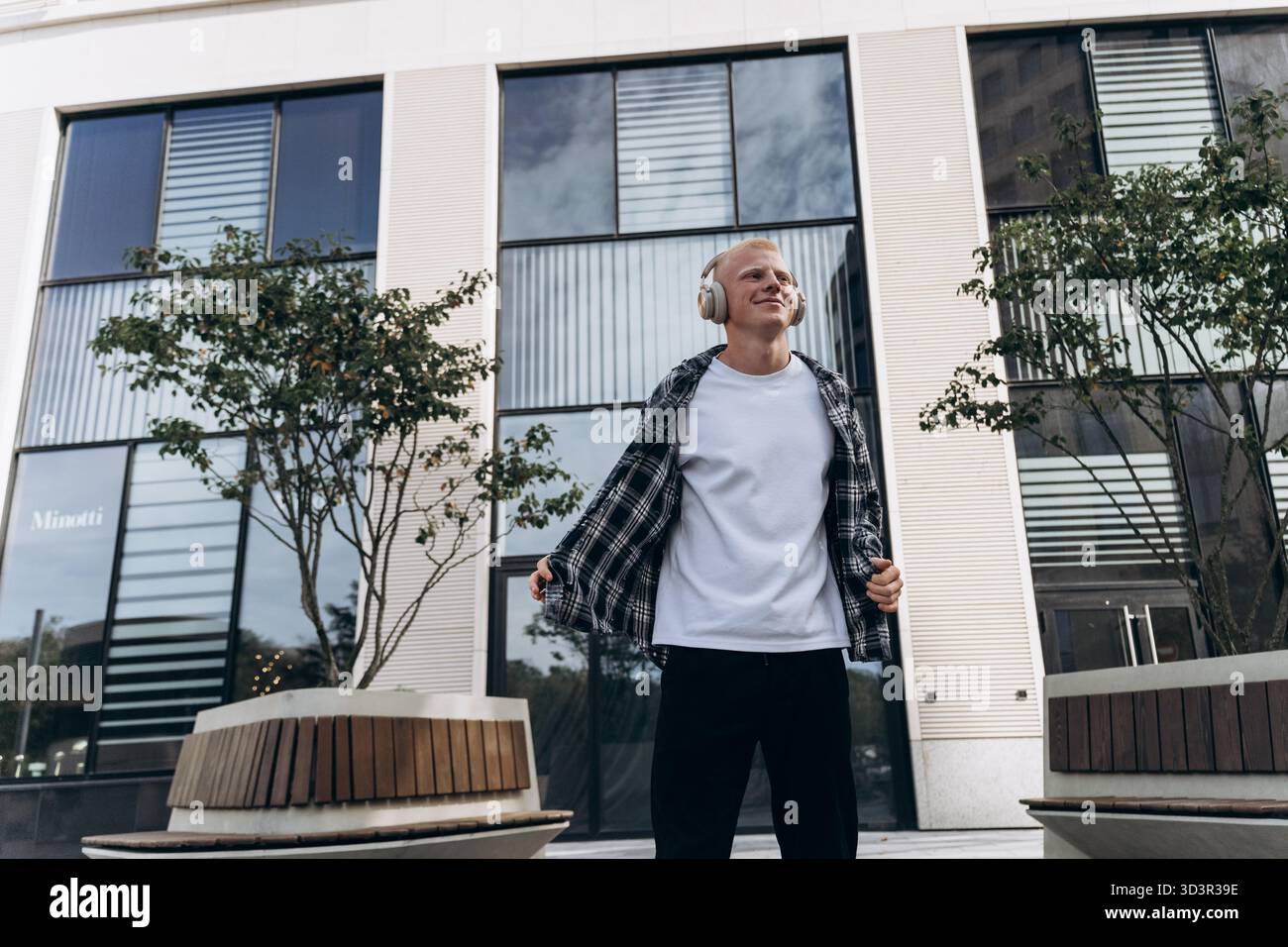 Ein junger Mann mit Albino-Auftritt lächelt glückselig, während er in einer urbanen Umgebung Musik über Kopfhörer hört, und zeigt ein entspanntes und Stockfoto