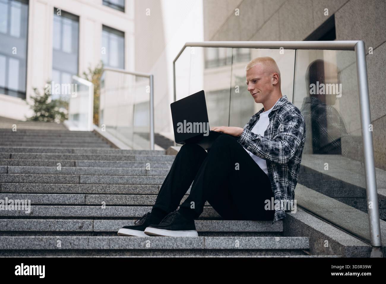 Ein junger Mann mit Albino-Aussehen sitzt auf städtischen Stufen, während er an seinem Laptop arbeitet. Er trägt legere Kleidung und die Szene spiegelt einen friedlichen Prod wider Stockfoto