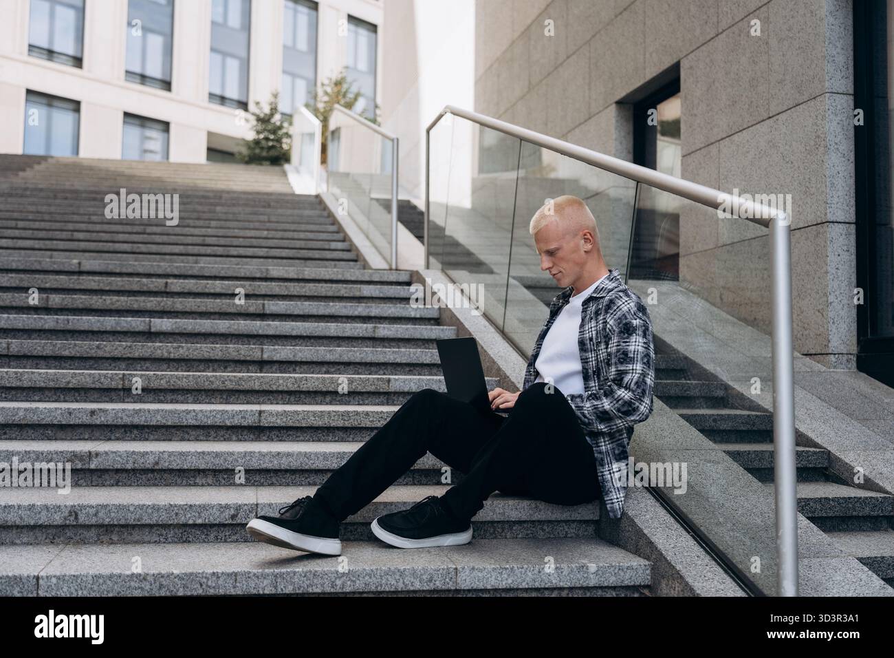 Ein junger Mann mit Albino-Aussehen sitzt auf städtischen Stufen, während er an seinem Laptop arbeitet. Er trägt legere Kleidung und die Szene spiegelt einen friedlichen Prod wider Stockfoto