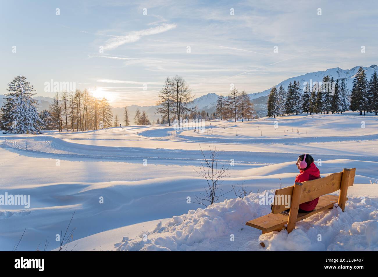 Eine ruhige Winterszene in den Alpen mit schneebedeckten Feldern, die sich im warmen Glanz eines Sonnenuntergangs sonnen. Eine Frau genießt die ruhige Aussicht aus einem Holz Stockfoto