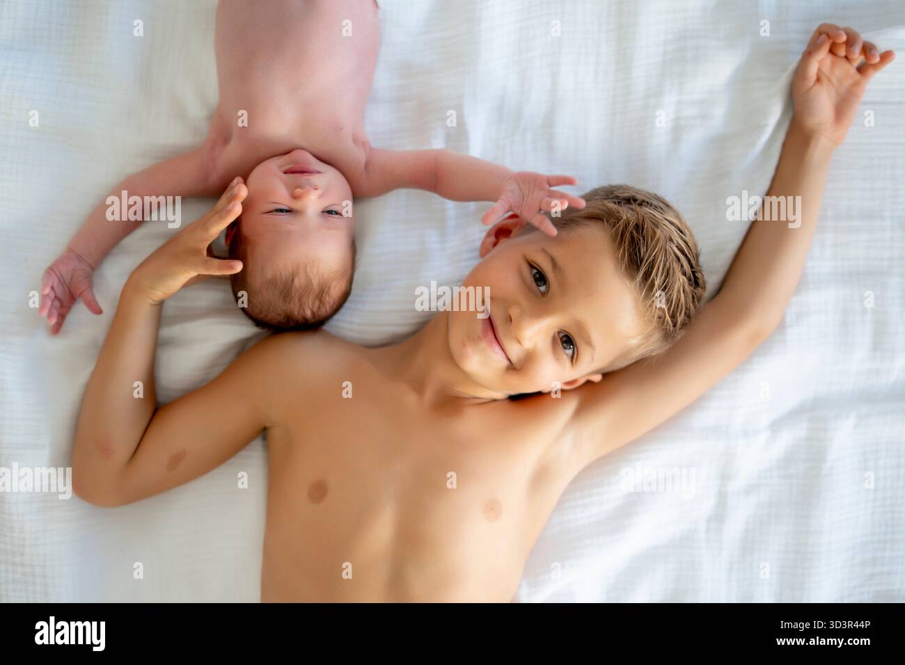 Zwei Geschwister liegen zusammen auf weißen Bettlaken und teilen einen freudigen Moment. Der ältere Bruder lächelt, während das Baby bequem ausruht, und hebt das hervor Stockfoto