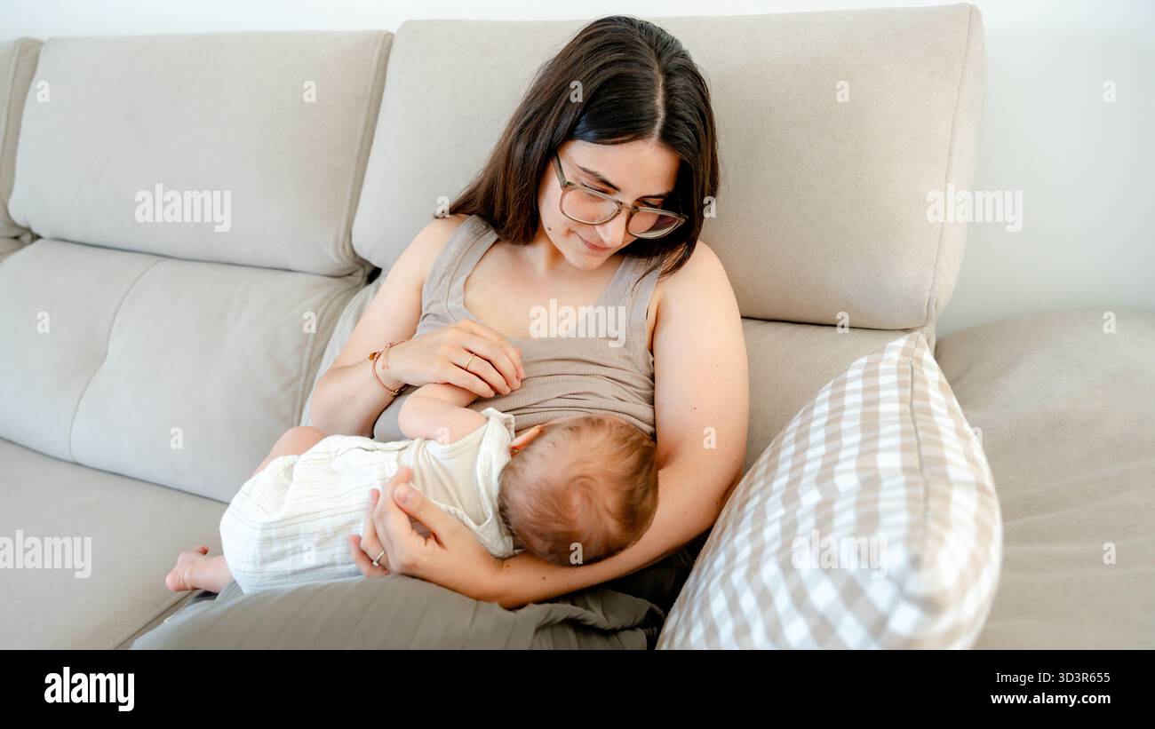 Ein pflegender Moment, in dem eine Mutter ihr 3 Monate altes Baby auf einem bequemen Sofa liebevoll stillt und die Verbundenheit und Zärtlichkeit der Eltern unterstreicht Stockfoto
