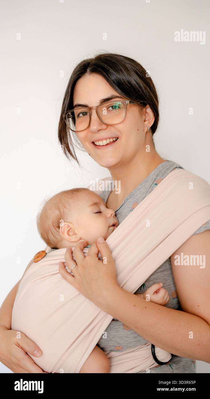 Eine Mutter lächelt warm, während sie ihr schlafendes Baby in einer weichen Hülle hüllt. Der intime Moment fängt die starke Verbundenheit und die pflegende Umgebung in sich ein Stockfoto