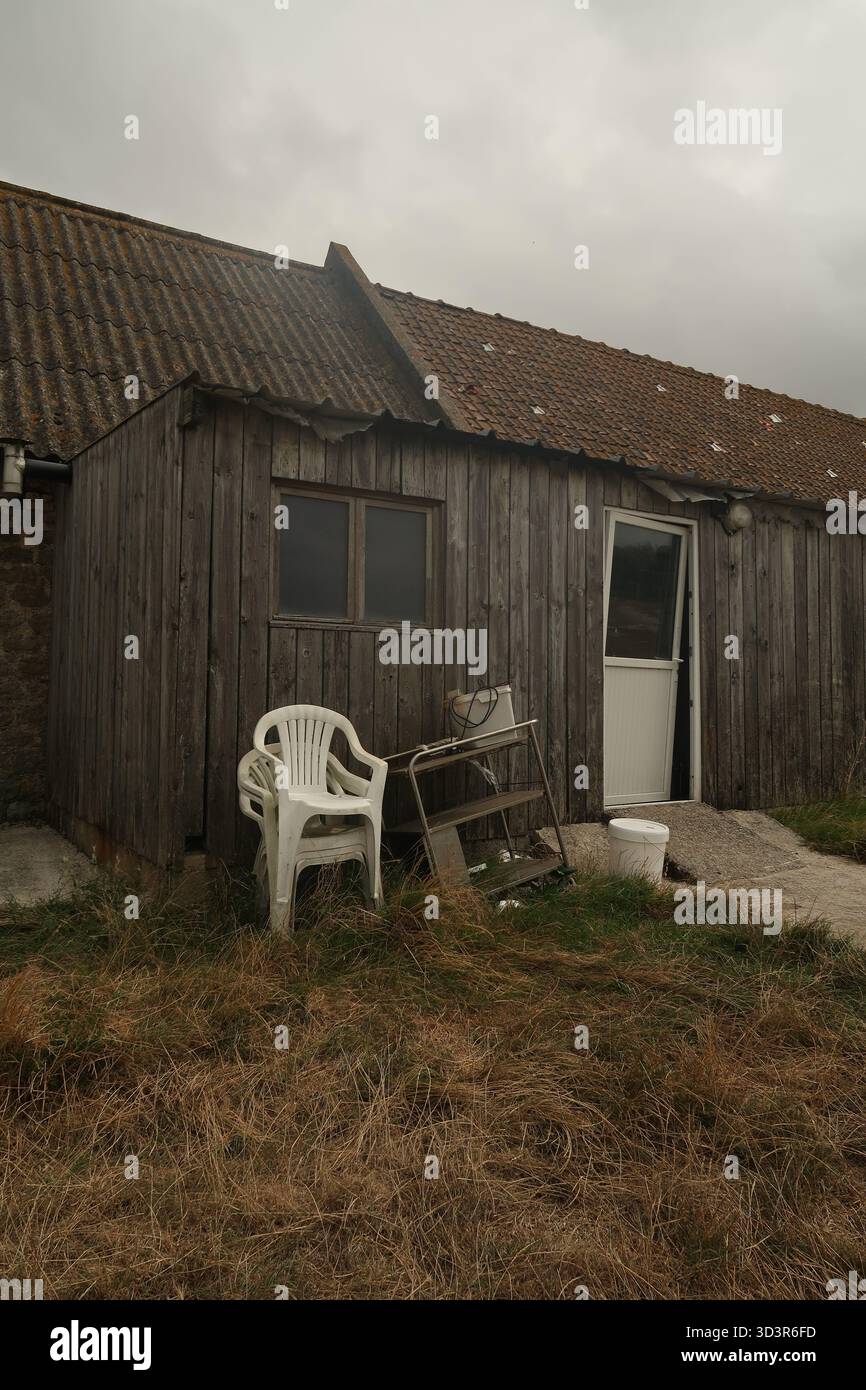 Eine verwitterte Holzscheune mit einem alten Stuhl und Ausrüstung draußen, vor einem bewölkten Himmel in der ländlichen Landschaft Nordfrankreichs, verkörpert Rusti Stockfoto