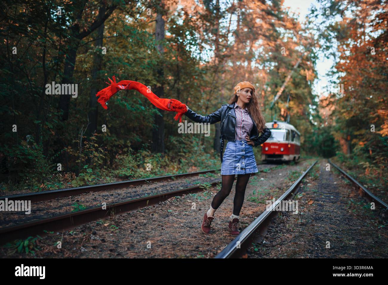 Eine Frau in Herbstkleidung trägt einen fließenden roten Schal, während sie auf Eisenbahngleisen steht, inmitten eines Waldes, der mit lebhaftem Herbstlaub gefüllt ist, was an einen erinnert Stockfoto