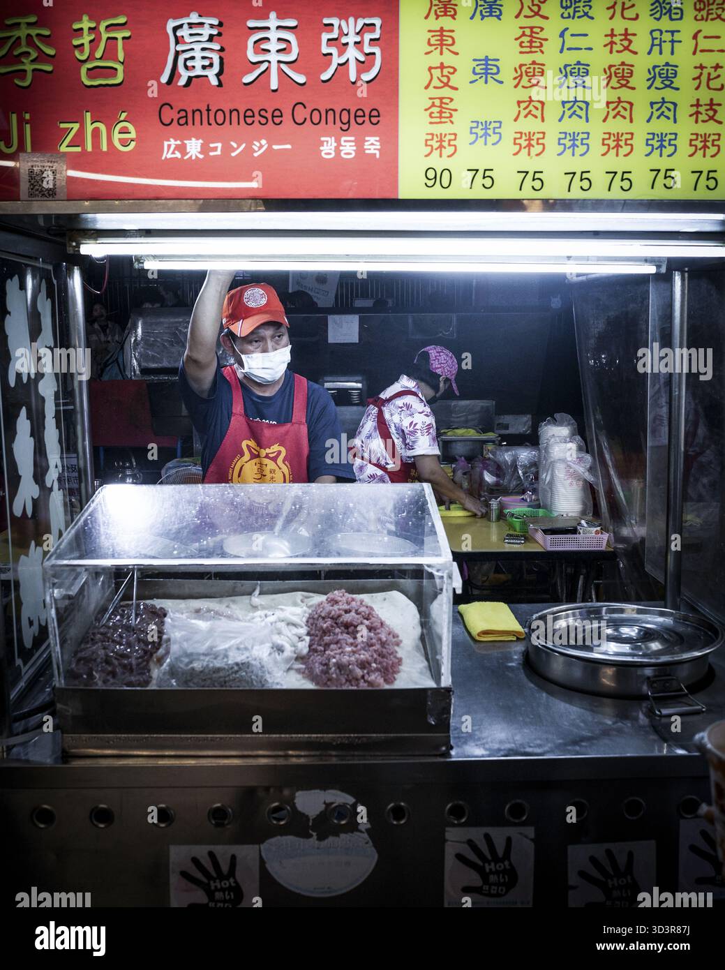 Taipeh, Taiwan - 20. Mai 2023: Blick auf einen pulsierenden kantonesischen Congee-Lebensmittelstand, wo ein maskierter Verkäufer in einer roten Schürze unter der hellen Grippe Gerichte zubereitet Stockfoto
