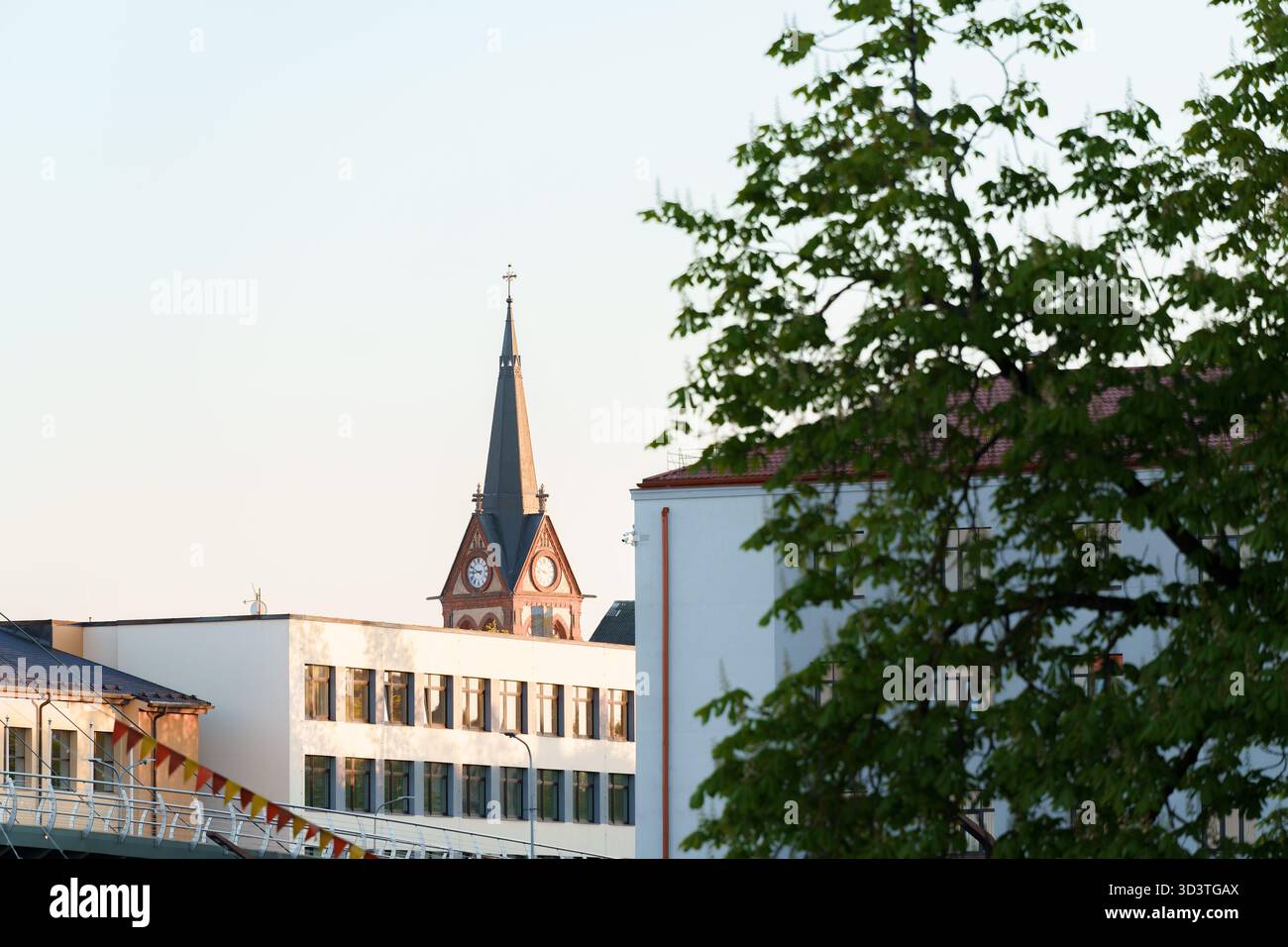 Sehr nah an Kirchtürmen mit Uhren in einer städtischen Umgebung. Uhren sichtbar auf dem Turm, Bäume und Stadtgebäude im Vordergrund, Jelgava, Stockfoto