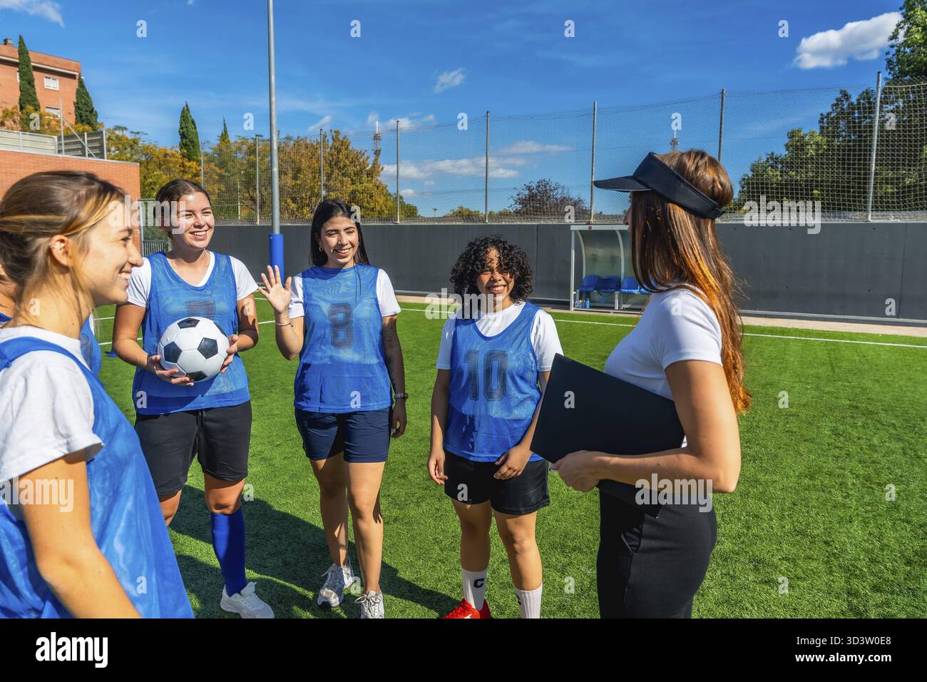 Fußballspielerinnen in blauen Pinnies hören aufmerksam zu, wie ihre Trainerin Strategien erklärt und die Mannschaft auf einem sonnigen Kunstgras führt Stockfoto