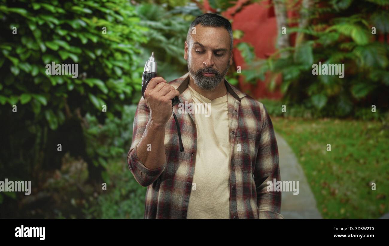 Mann, der einen elektrischen Bohrer in der rechten Hand hält, auf einem Straßenweg, der von grünem Parklaub eingerahmt ist, Bart und Schreinerflanell sichtbar ist; entschlossenes Handwerkskunst Stockfoto
