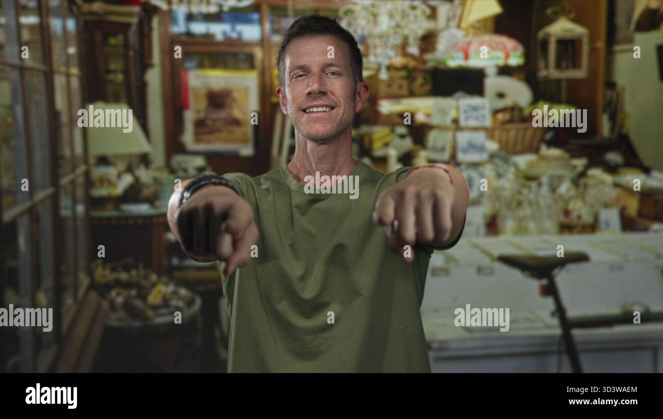 Der Mann zeigt mit dem Finger auf die Kamera im Ladengebäude, während er lächelt und ein grünes Hemd und Armbänder trägt; Willkommen. Stockfoto