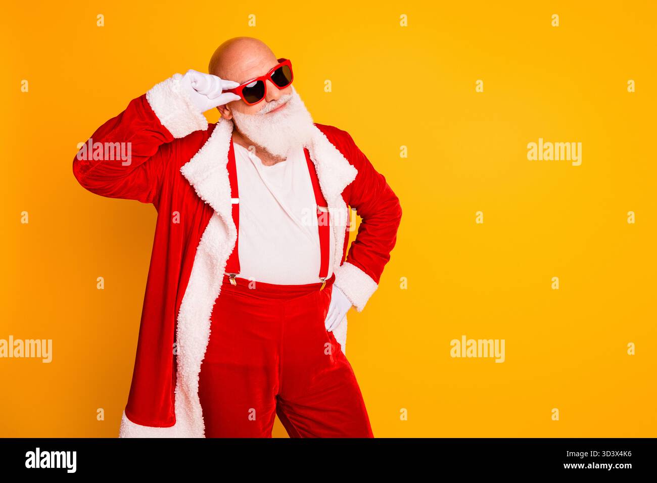 Weihnachtsmann in rotem Kostüm mit weißem Pelzbesatz in gelbem Studio sorgt mit selbstbewusstem Lächeln im Retro-Stil für festliche Weihnachtsstimmung Stockfoto