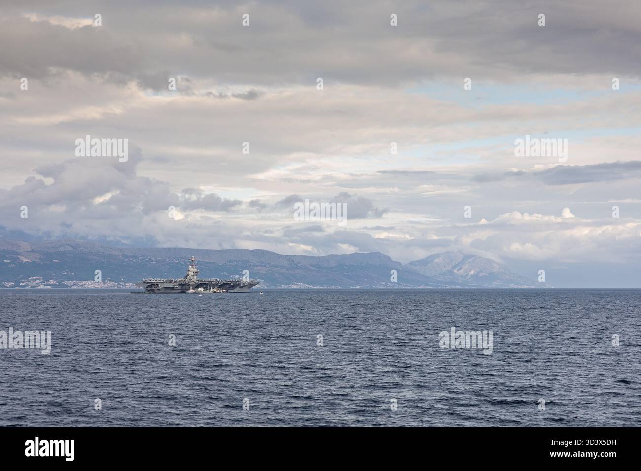 Ein riesiger Flugzeugträger segelt über ruhiges Wasser mit einer zerklüfteten Küste und weit entfernten Bergen unter einem grauen, bewölkten Himmel. Eine mächtige maritime sce Stockfoto