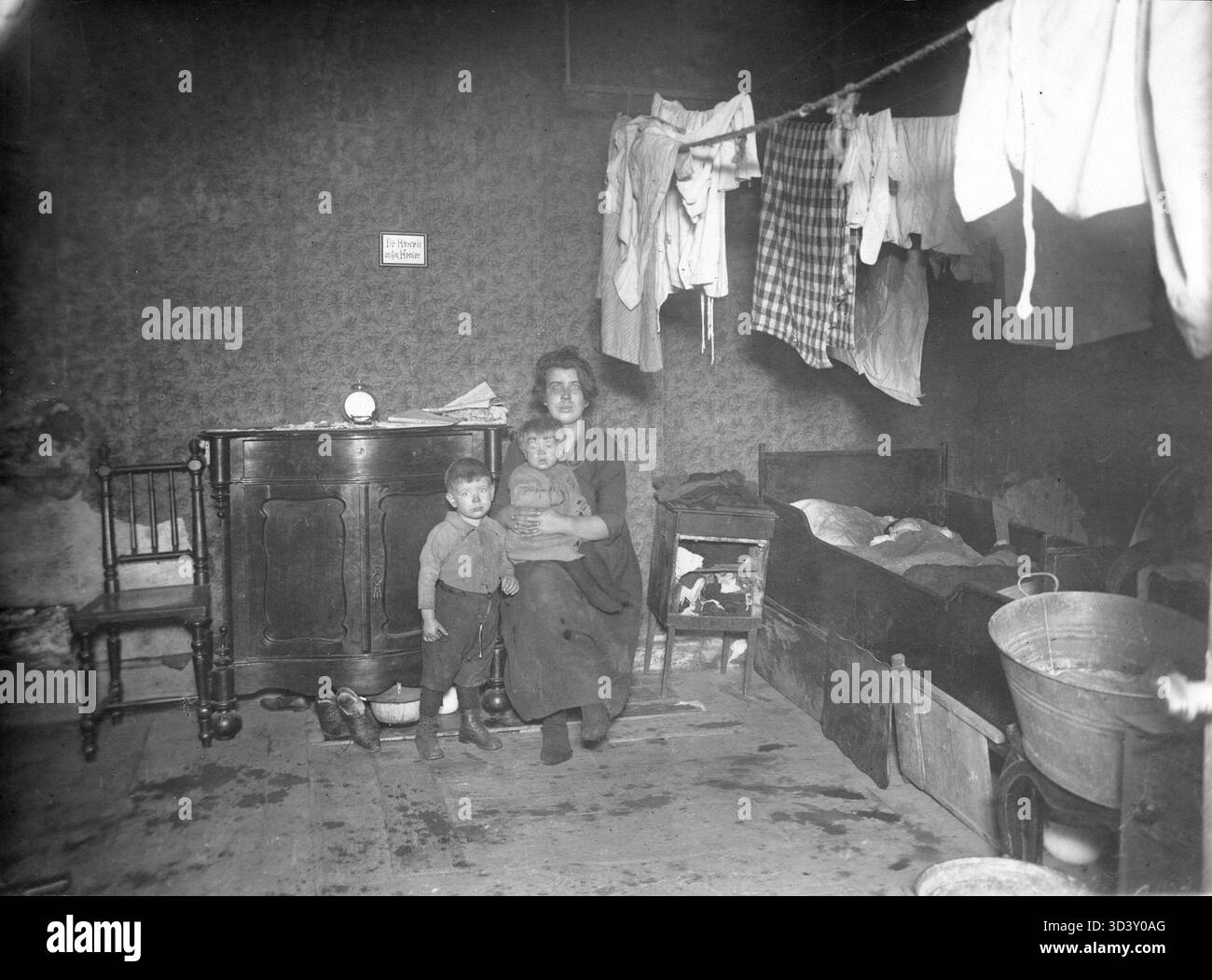 1928 werden eine Frau und Kinder in ihrem Haus auf Kleeresloot 2, Amsterdam, dargestellt, die unter verarmten Bedingungen leben. Das Zimmer verfügt über ein Bett, ein Waschbecken, ein Waschbecken und Kleidung, die an den Washlinen darüber hängen. Ein gerahmtes Sprichwort: "Der Herr ist mein Hirte", hängt an der Wand. Stockfoto
