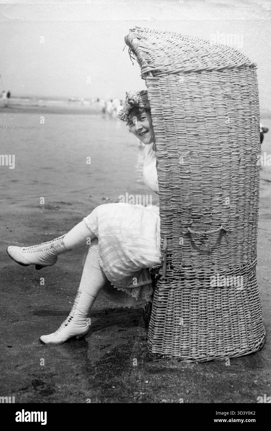 Im Jahr 1919 schaut sich eine Frau in weiß, einschließlich weißer Strümpfe und Schnürschuhe, verspielt von einem Korbstuhl aus um. Dies fängt einen Moment des Strandlebens im frühen 20. Jahrhundert ein. Stockfoto