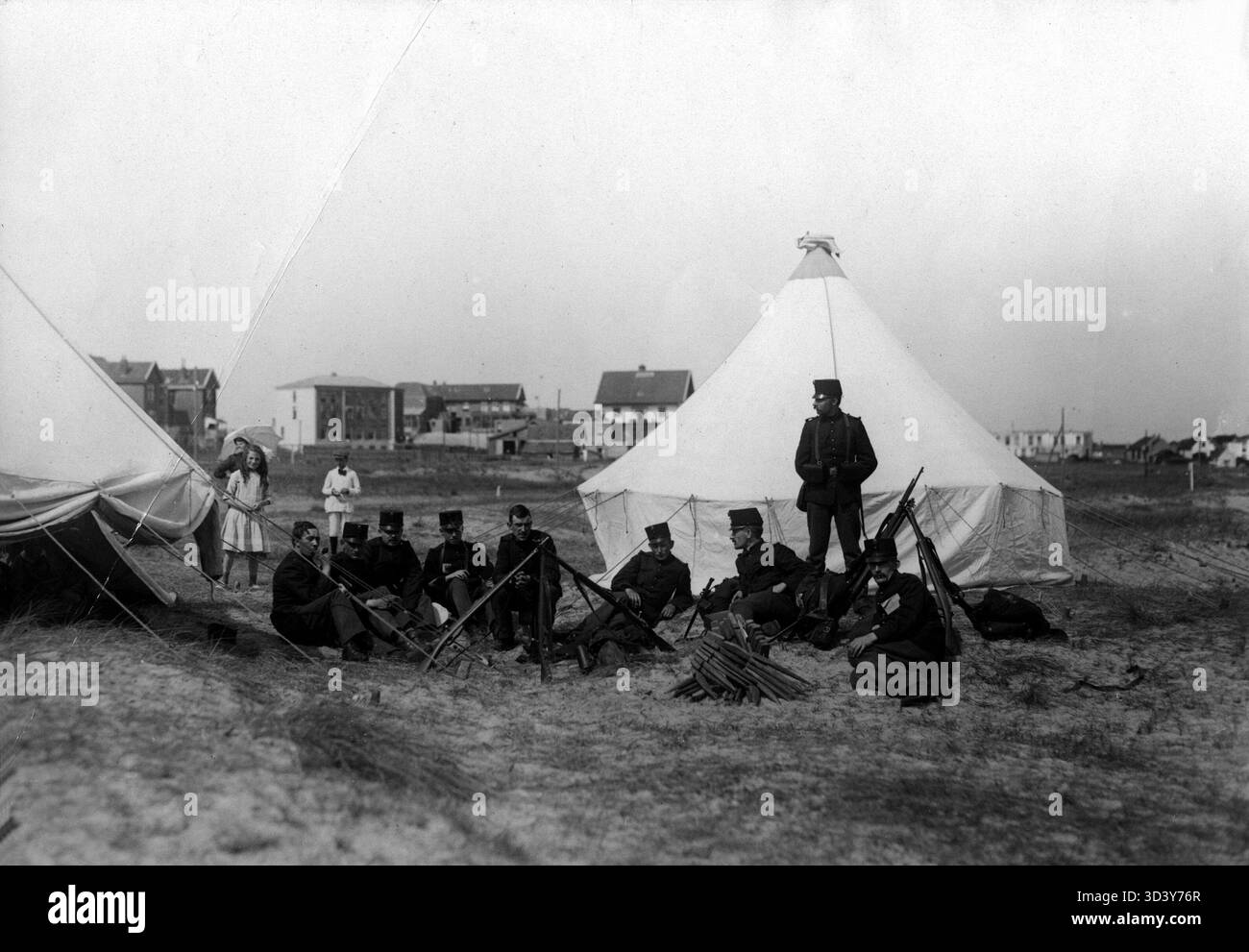 Während der Mobilisierung 1914 sind niederländische Soldaten an der Küste stationiert. Soldaten, bewaffnet mit Gewehren, sitzen an ihren Zelten in den Dünen, während Kinder beobachten. Im Hintergrund ist ein Badeort zu sehen. Stockfoto
