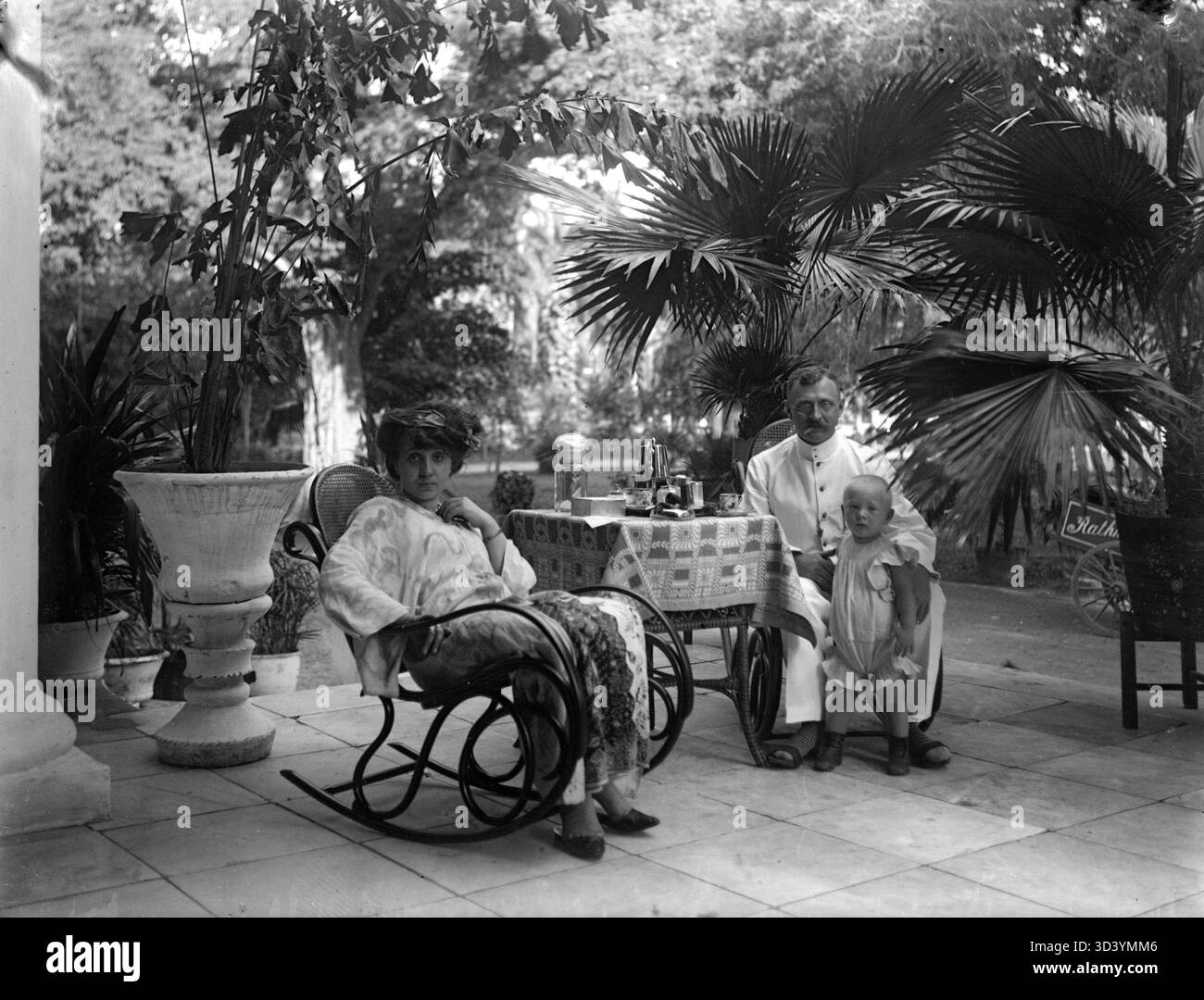 Johannes Maas, Melina Maas-Elzinga und ihr Sohn Bob sitzen auf Thonet Schaukelstühlen an einem Teetisch im Garten ihres Hauses in Batavia. Im Hintergrund, Palmen in Töpfen und ein Wagen mit der Inschrift Rath... Von Rathkamp & Co. Um 1910. Stockfoto