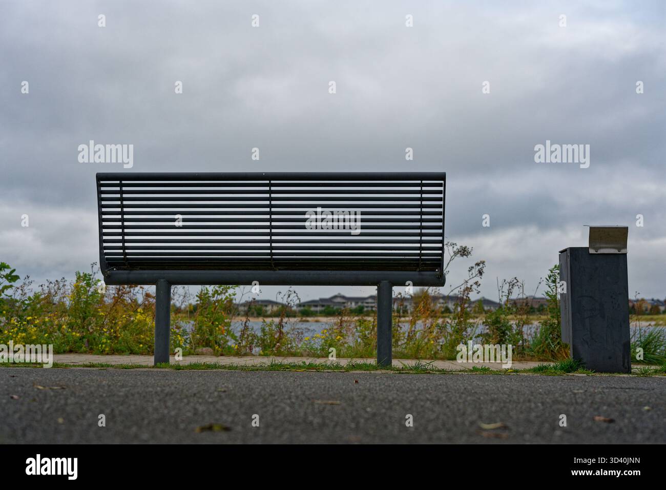 Eine hölzerne Bank blickt auf ruhiges Wasser mit einem kleinen Abfalleimer daneben, kühle Luft bewegt sich entlang der Küste und das einfache Paar bietet eine ruhige Pause zum Ausruhen und Stockfoto