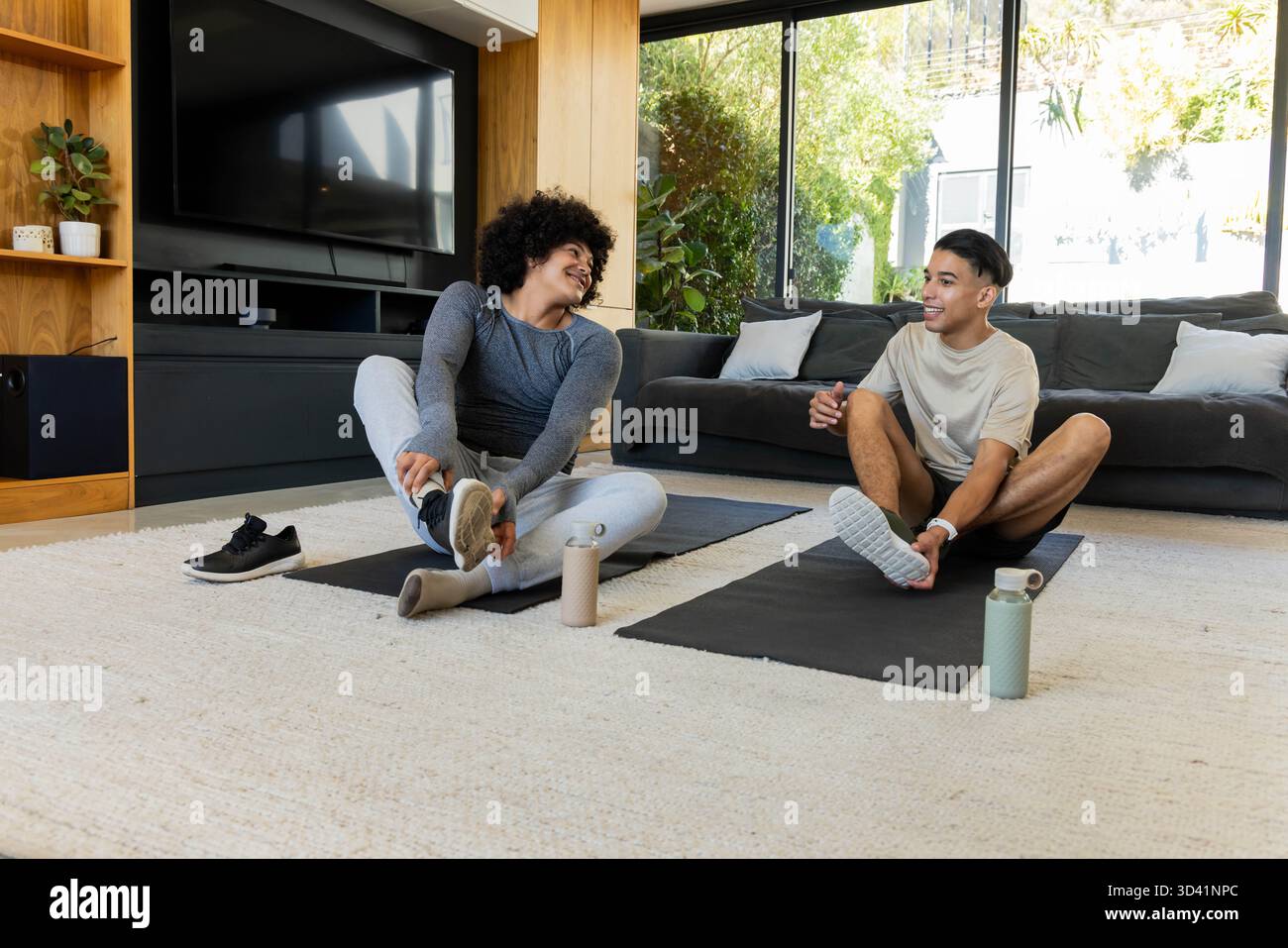 Verschiedene männliche Freunde dehnen und justieren Socken auf Yogamatten im Wohnzimmer mit Wasserflaschen Stockfoto