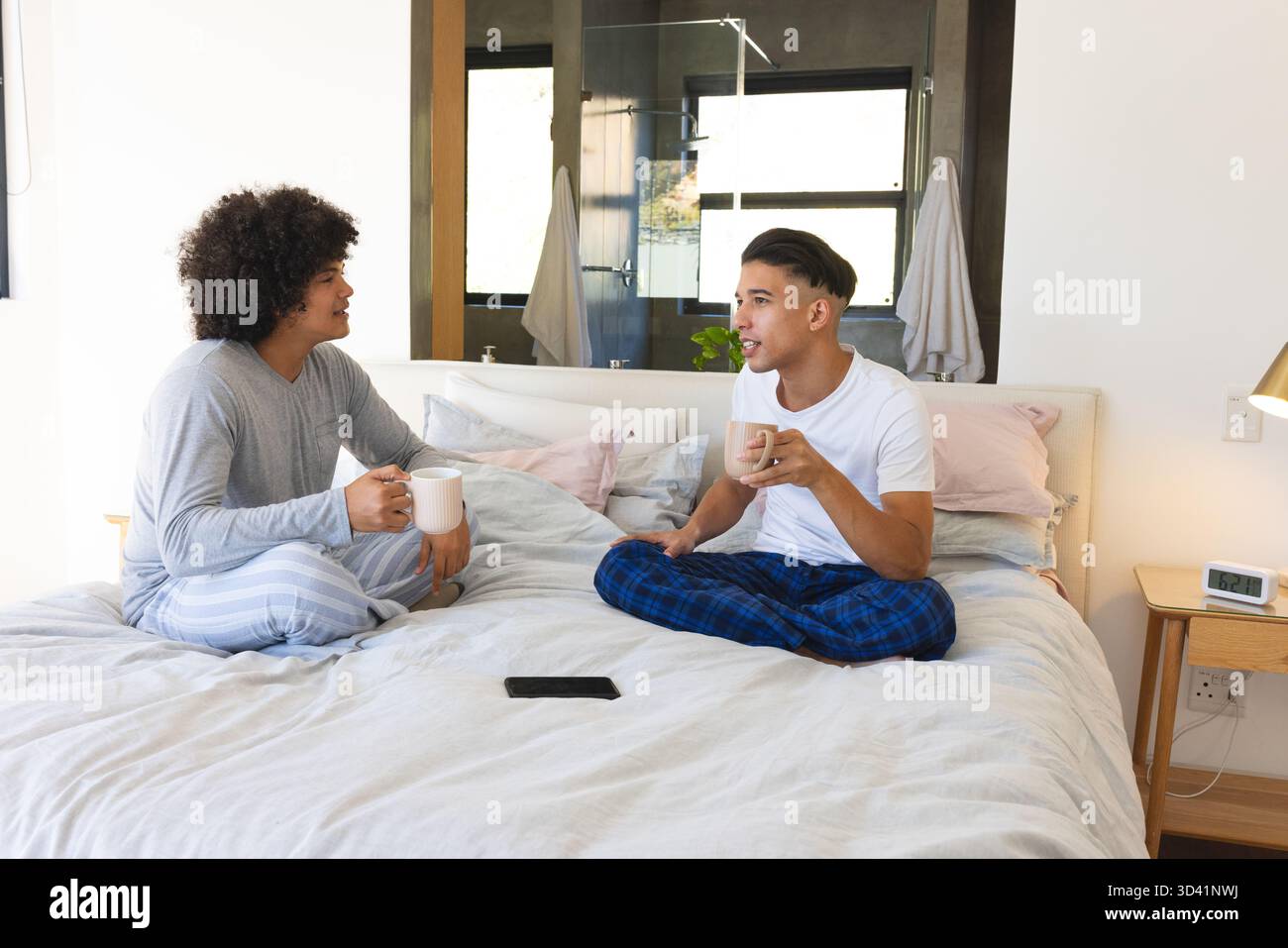 Unterhalten Sie sich mit verschiedenen männlichen Freunden auf dem Bett im Schlafzimmer, mit Kaffeetassen und Smartphone Stockfoto
