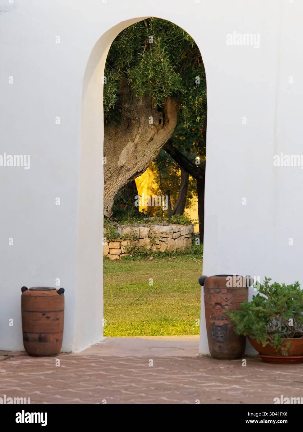 Eingang mit gewölbter weißer Wand, der üppigen Garten mit Steinmauer und Töpfen umrahmt Stockfoto