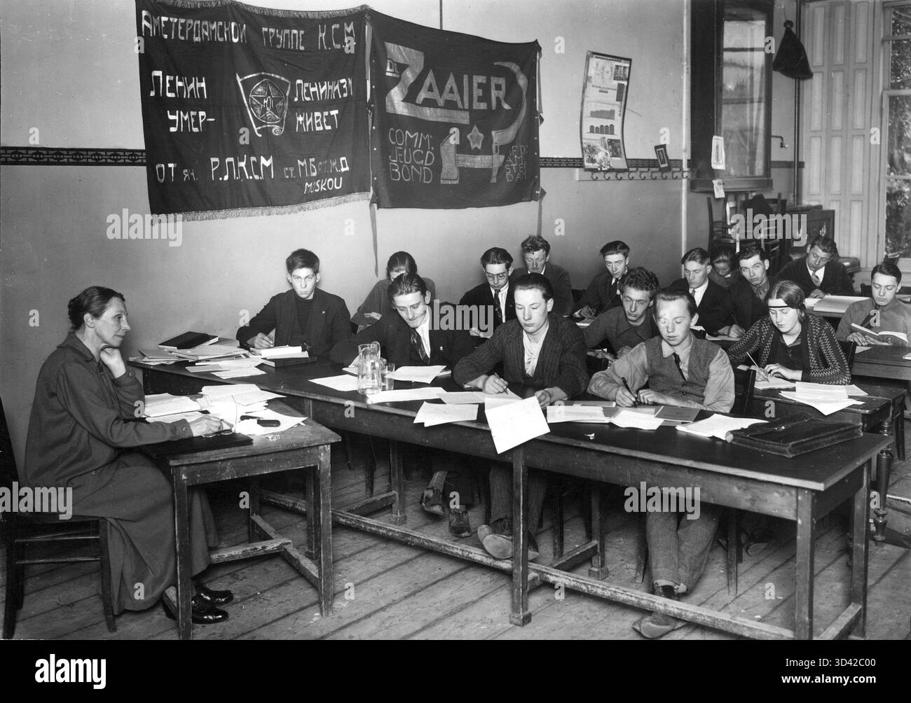 Foto von Henriette Roland Holst mit Mitgliedern der Kommunistischen Jugendliga de Zaaier in Amsterdam um 1926. Die Szene zeigt junge Menschen, die mit Büchern und Papieren an Tischen sitzen. An der Wand hängen Banner, darunter eines auf Russisch und ein anderes mit dem Hammer- und Sichelsymbol. Karten werden auch im Raum angezeigt. Stockfoto